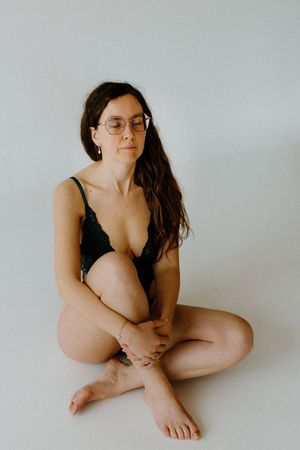 Woman in black lace bodysuit sitting cross-legged on white floor, wearing glasses with long dark wavy hair.