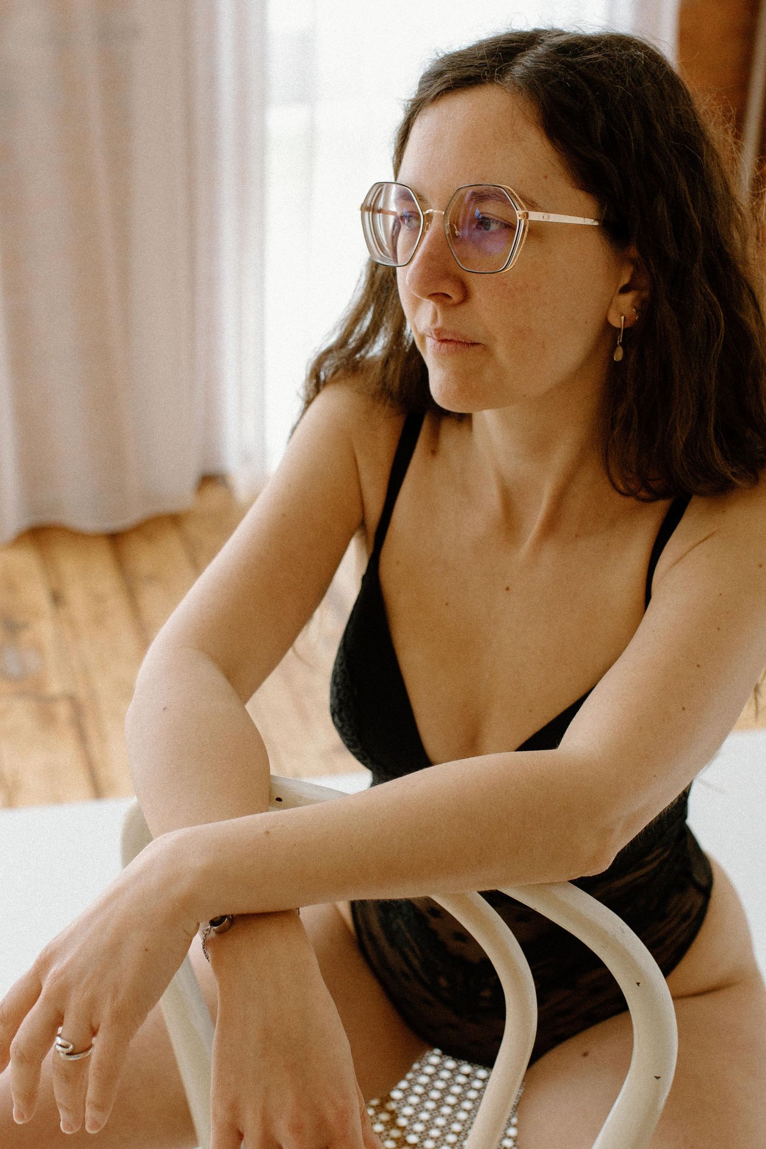 Woman in black lingerie sitting on a chair, wearing large clear-frame glasses, with long dark hair.