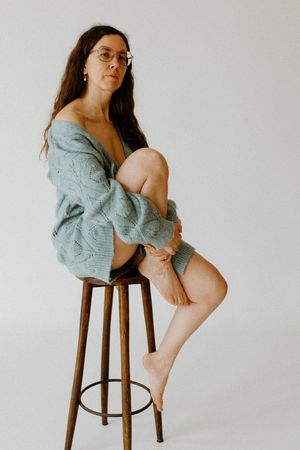 Woman in an oversized teal knit cardigan and shorts sitting on a wooden stool against a white background.