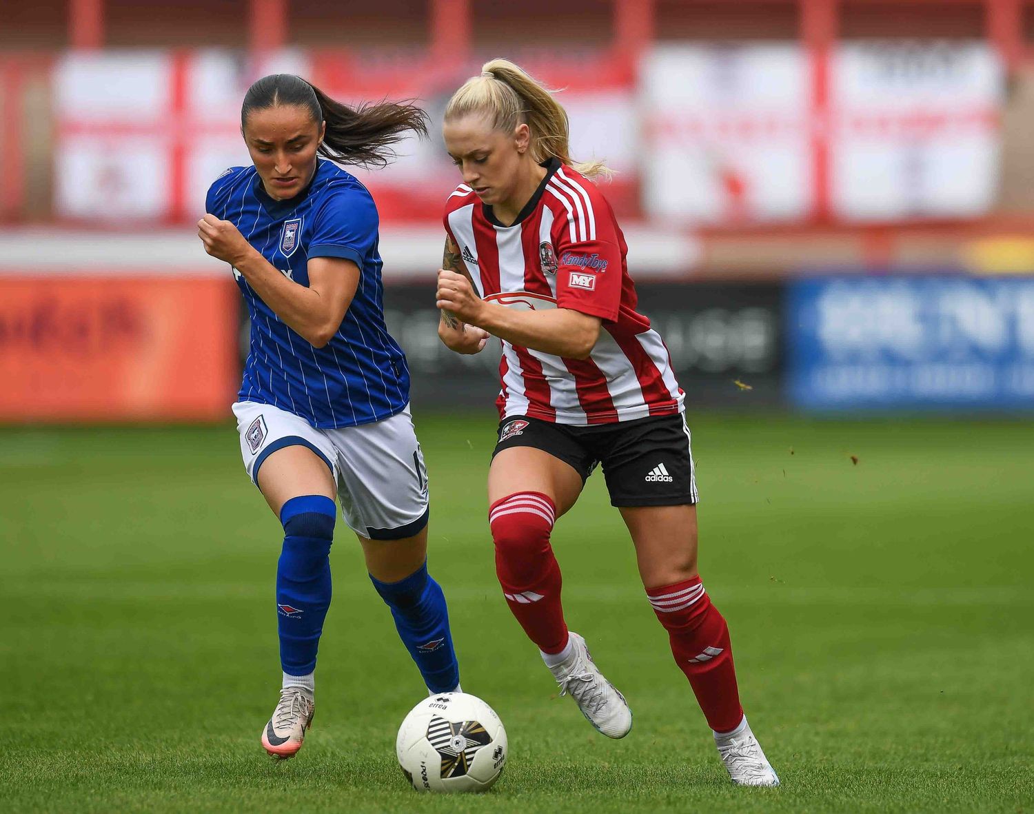 Two soccer players battle for possession of the ball during an intense match between teams in blue and red-striped jerseys.