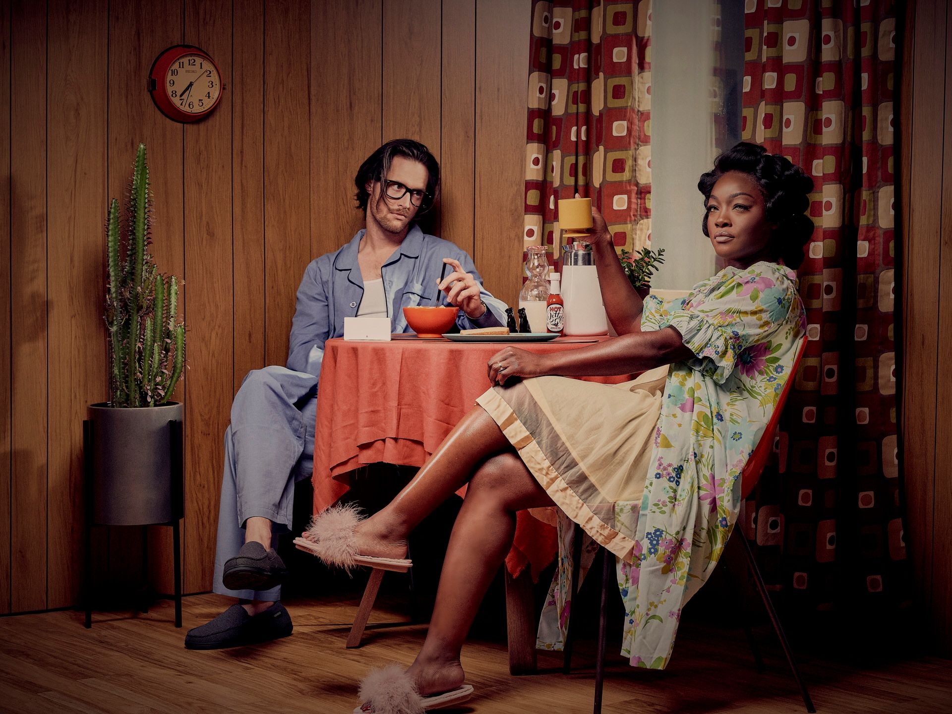 A retro-styled dinner scene with a couple sharing drinks at a small table with red tablecloth and floral curtains.