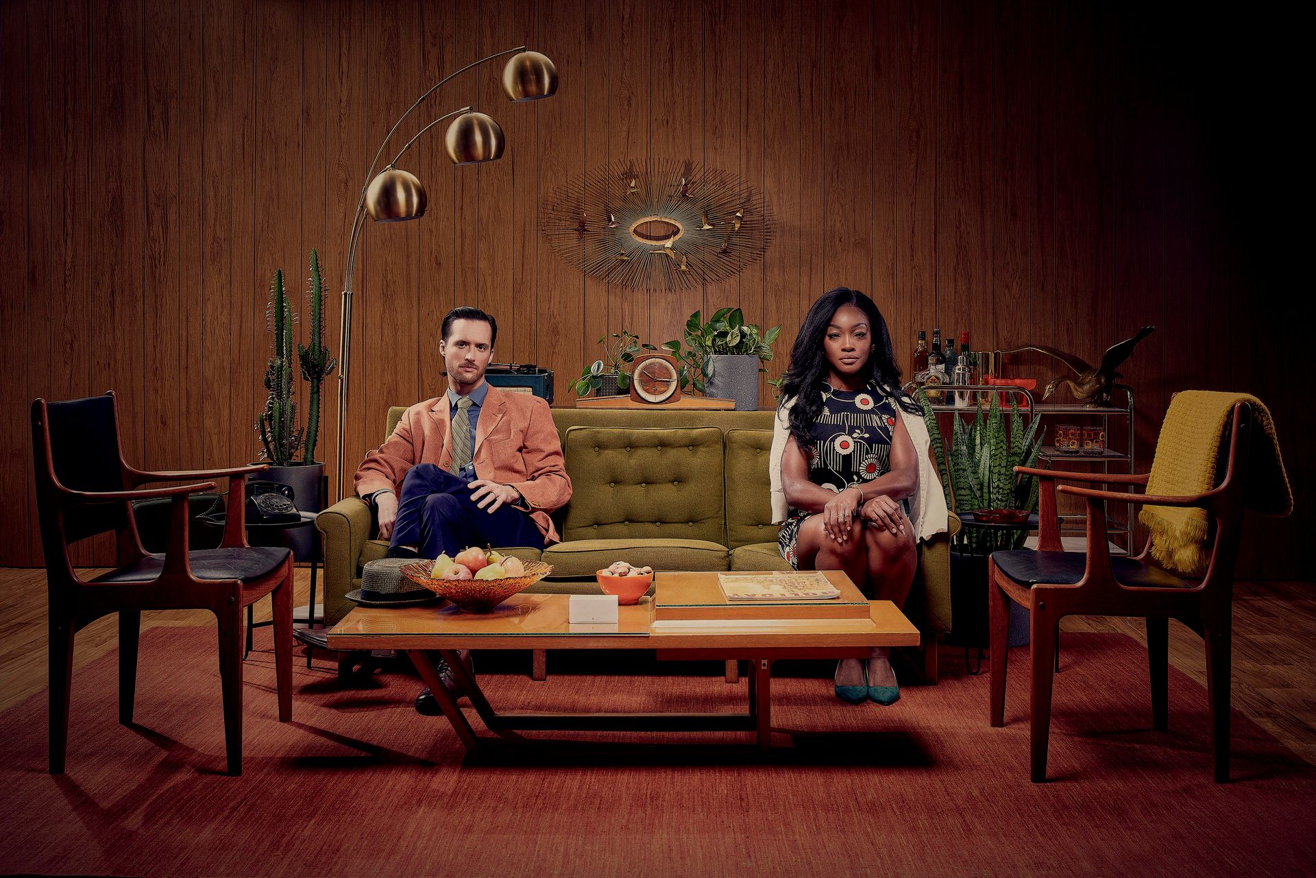 Two people sitting on a mustard yellow couch in a vintage-styled living room with wood paneling and retro decor.