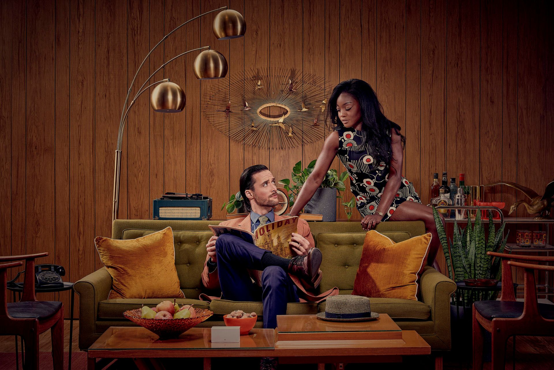A couple interacting on a vintage yellow couch with mid-century modern decor and wood paneled walls.