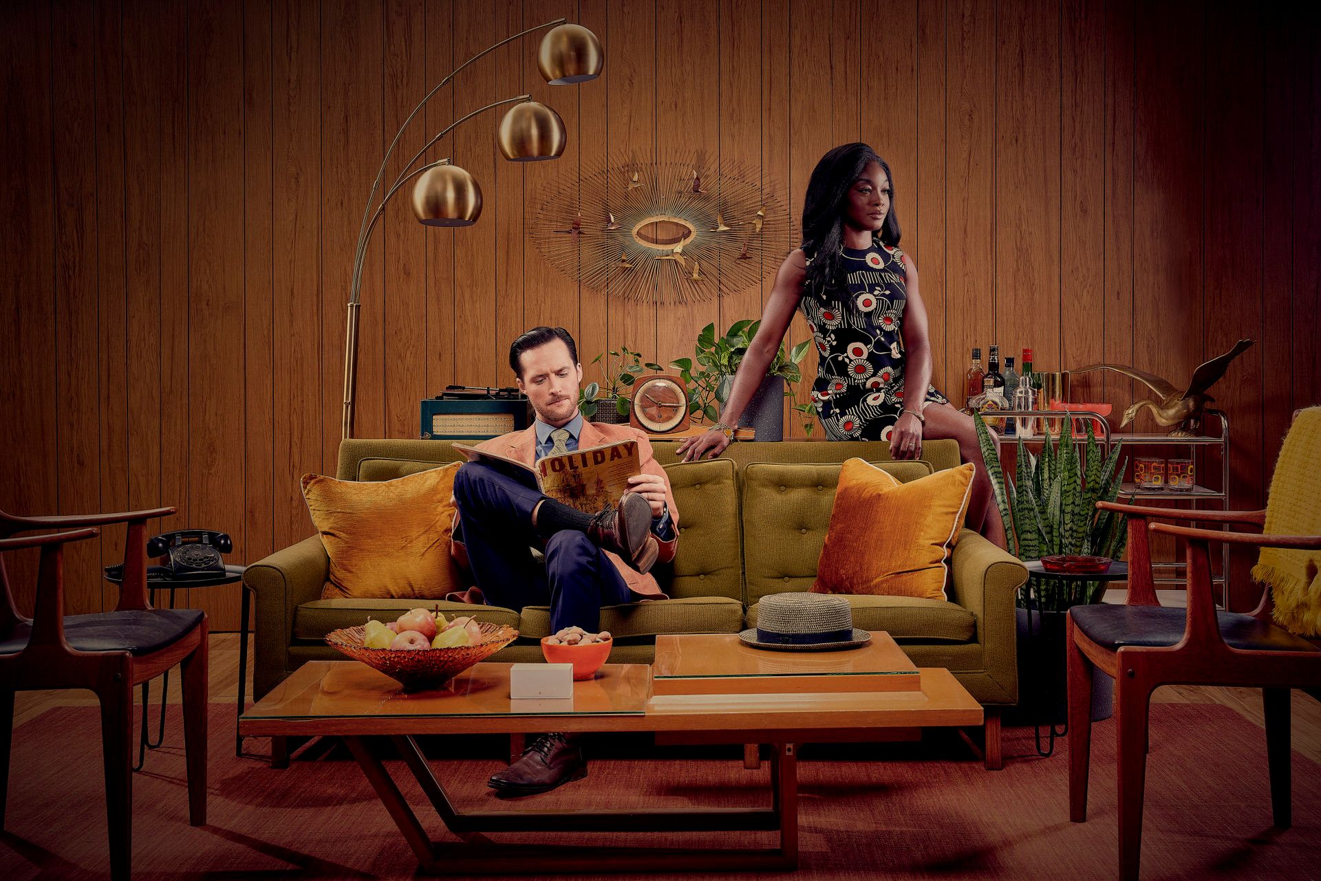 A mid-century modern living room scene with two people on a yellow couch surrounded by vintage decor.