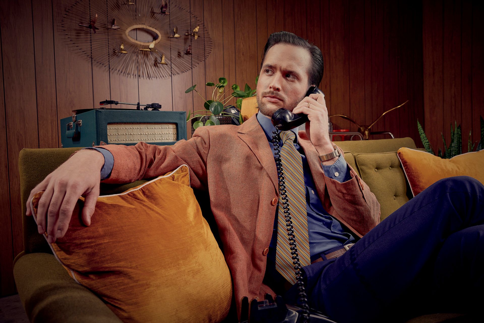 A person in a brown blazer lounging on a yellow couch while talking on a vintage telephone.