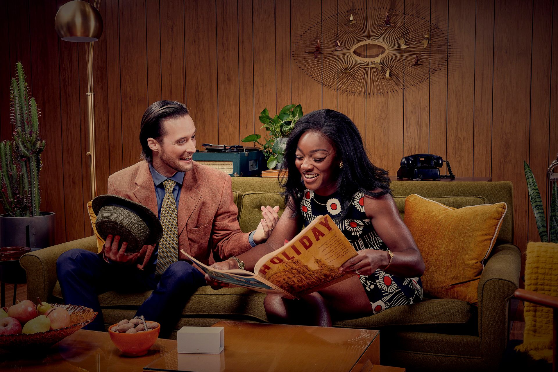 Two people sharing a casual moment on a yellow couch while reading in a retro-styled living room.