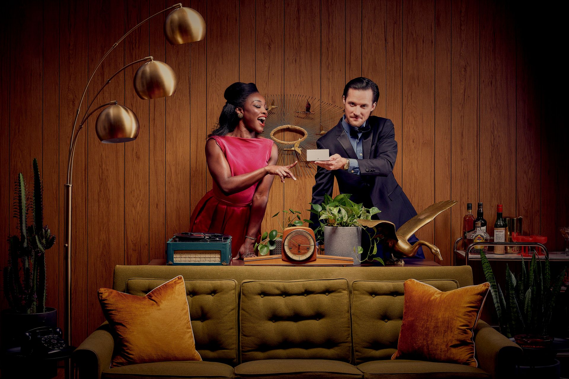 A playful scene with two people juggling oranges in front of a mid-century modern couch with yellow and orange pillows.
