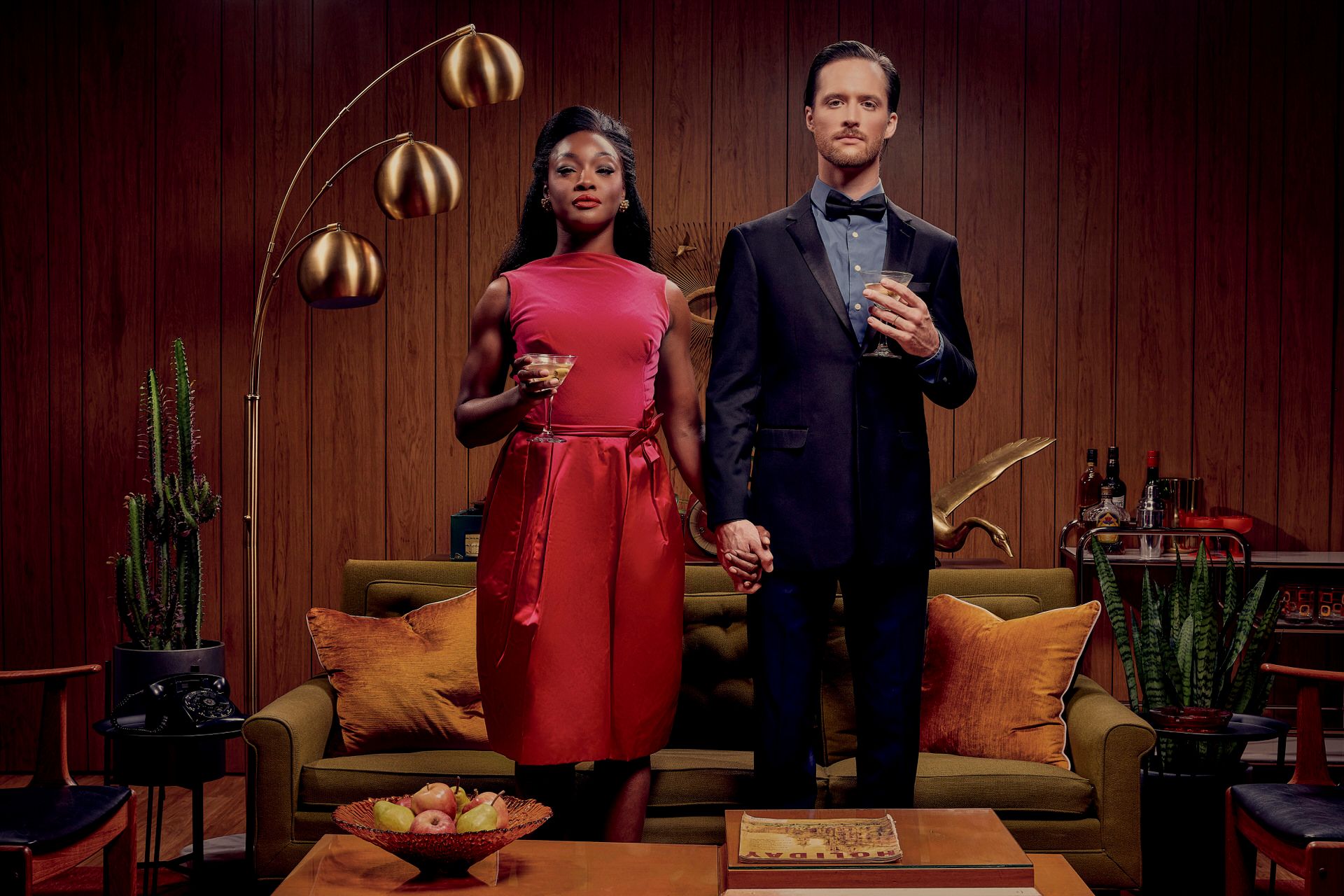 Stylish performers posing with oranges in a retro-decorated room with wood paneling and a green velvet couch.