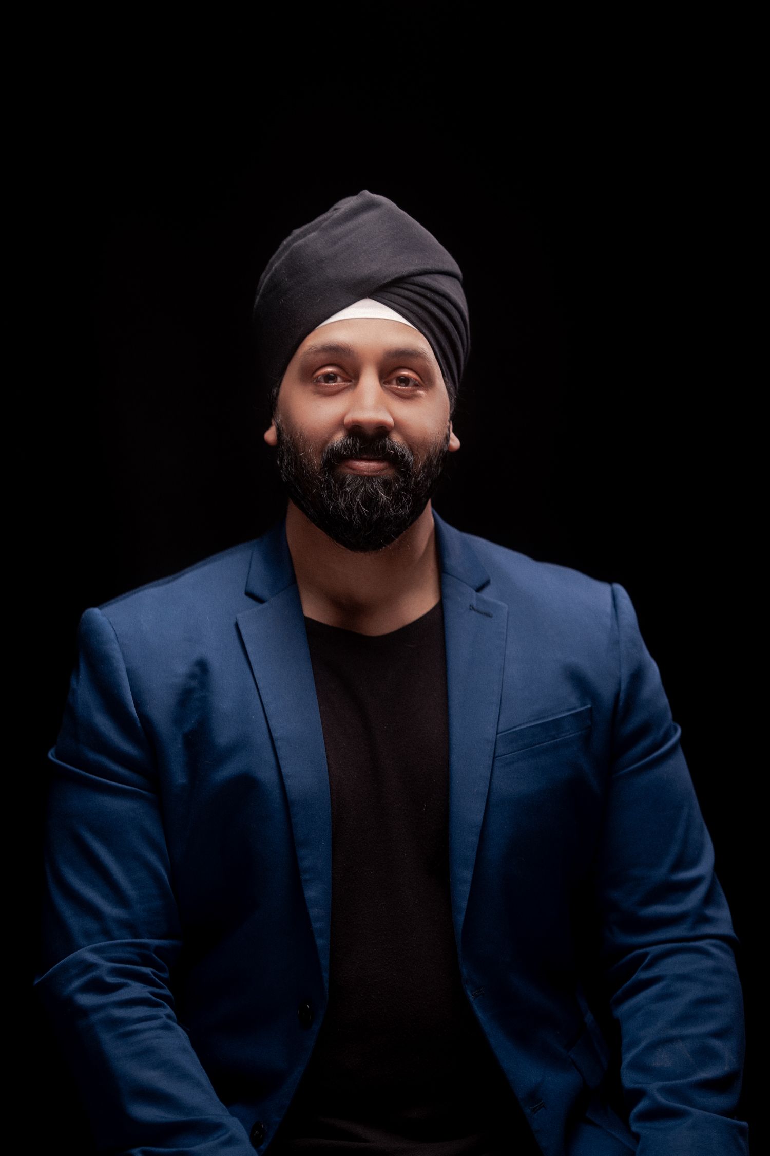 Professional studio portrait of someone in a black turban and navy blazer against dark background.