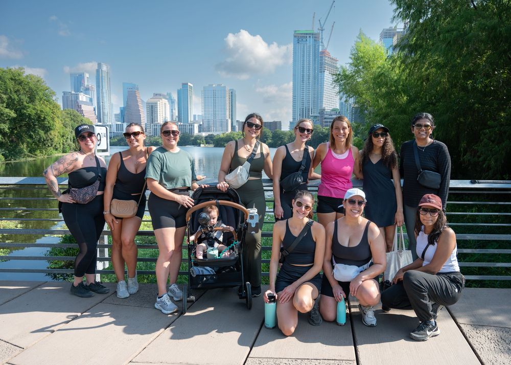 A group of women in athletic wear pose together on a bridge with a city skyline backdrop.