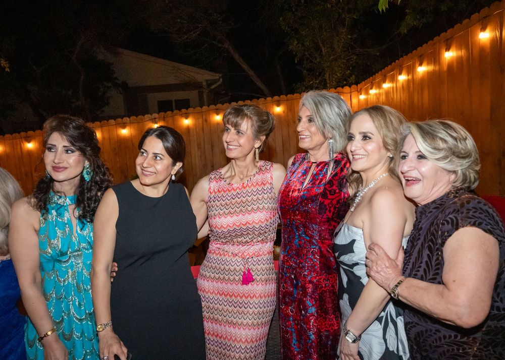 Six elegantly dressed women pose together at an outdoor evening celebration with string lights.