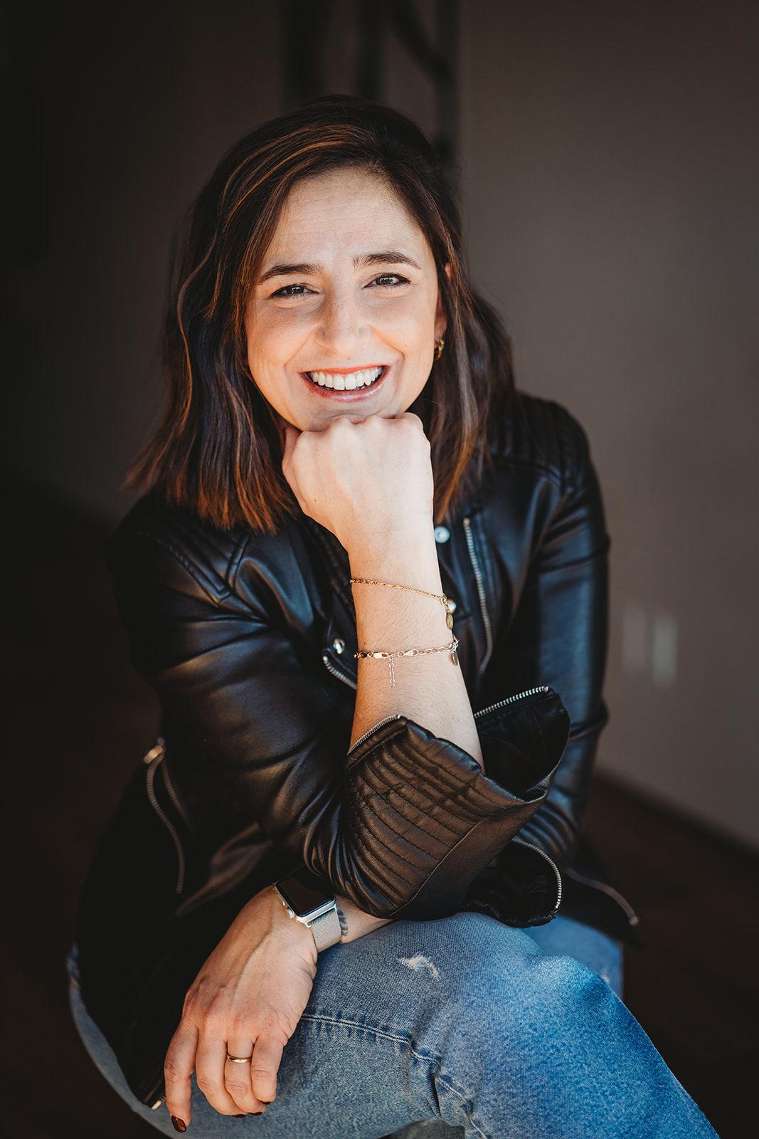 Portrait shot of someone in a leather jacket smiling while resting their chin on their hand.