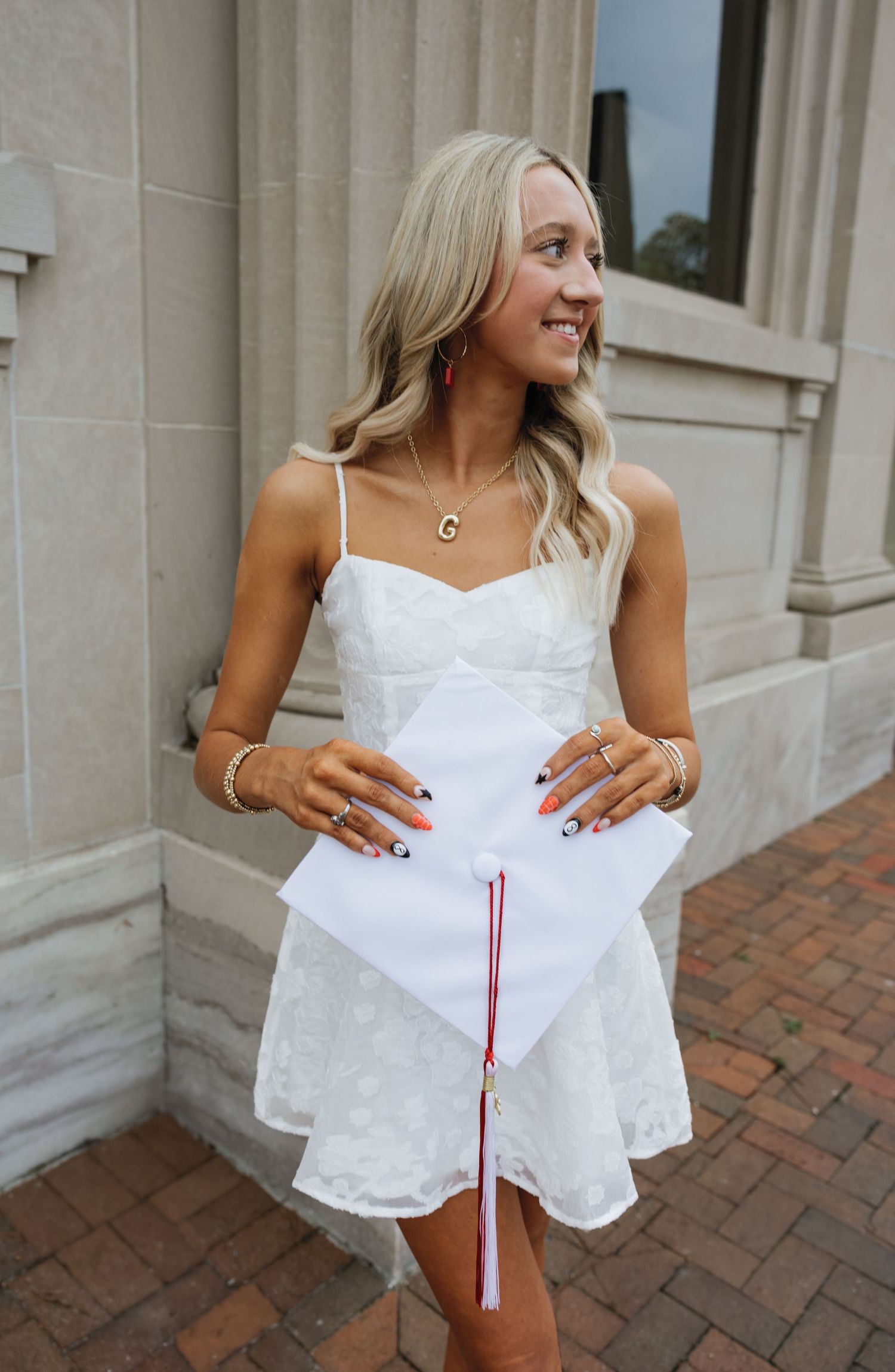 Woman in white graduation dress smiles while holding diploma against brick building.