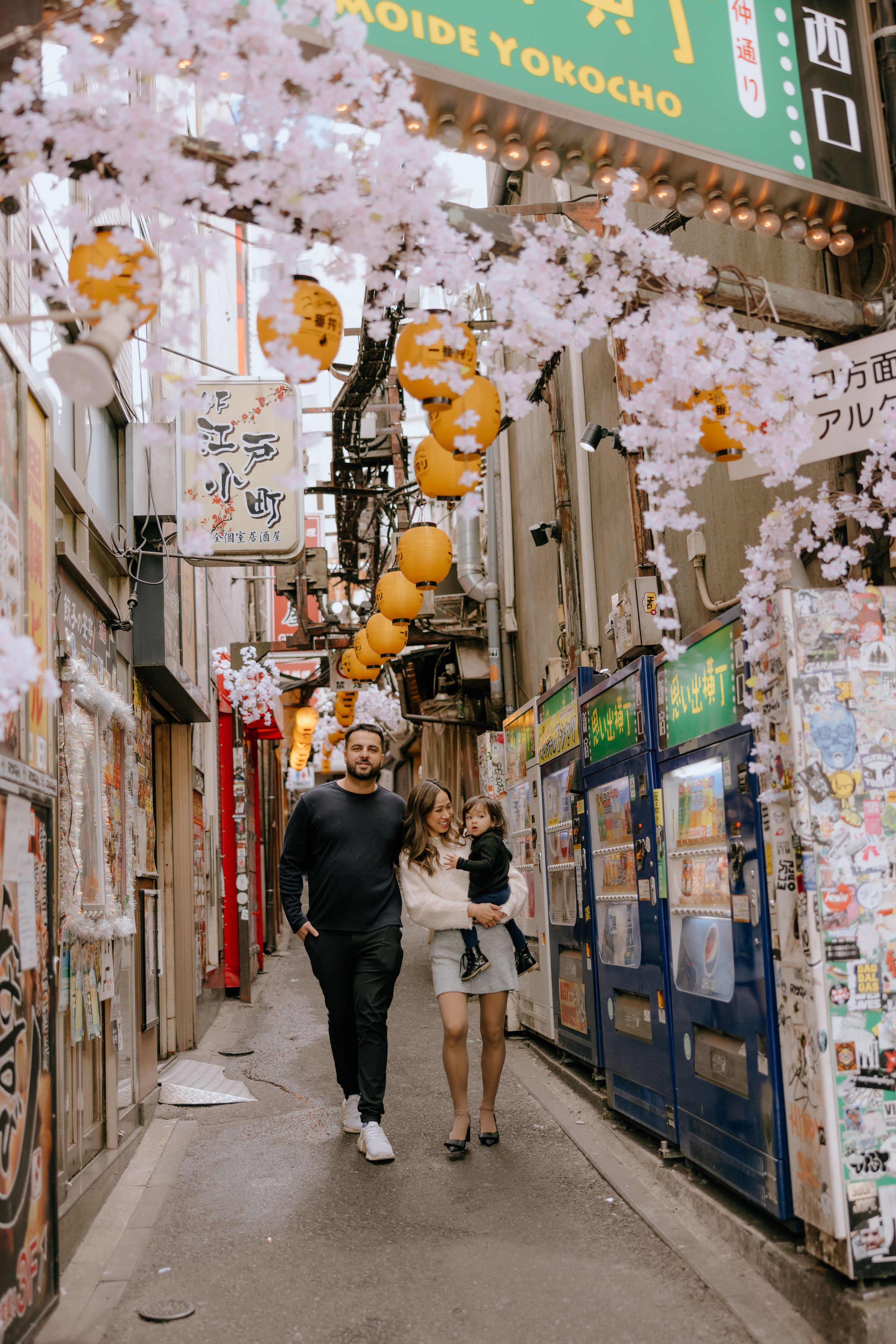 Narrow Japanese alleyway decorated with pink cherry blossoms and yellow lanterns in an urban shopping district.