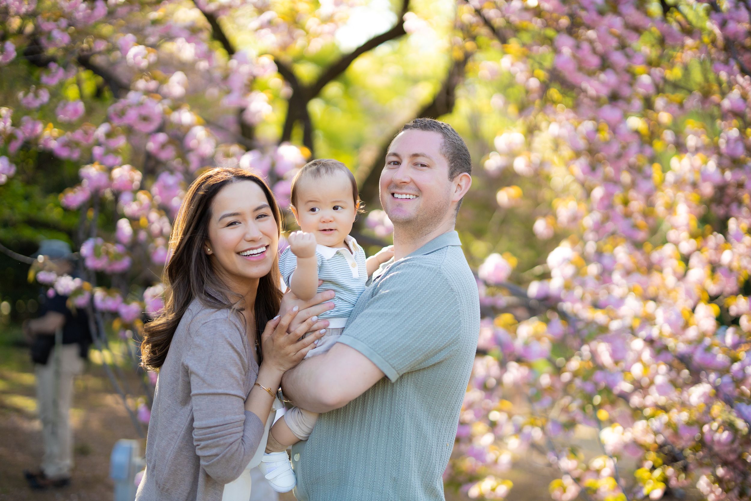 A family poses together under blooming pink cherry blossom trees in spring sunlight.