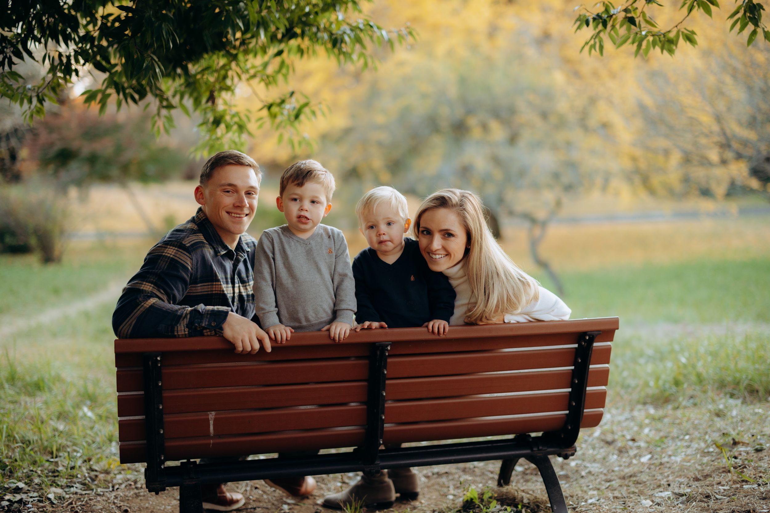 A family sits together on a wooden park bench during golden hour with autumn foliage in the background.