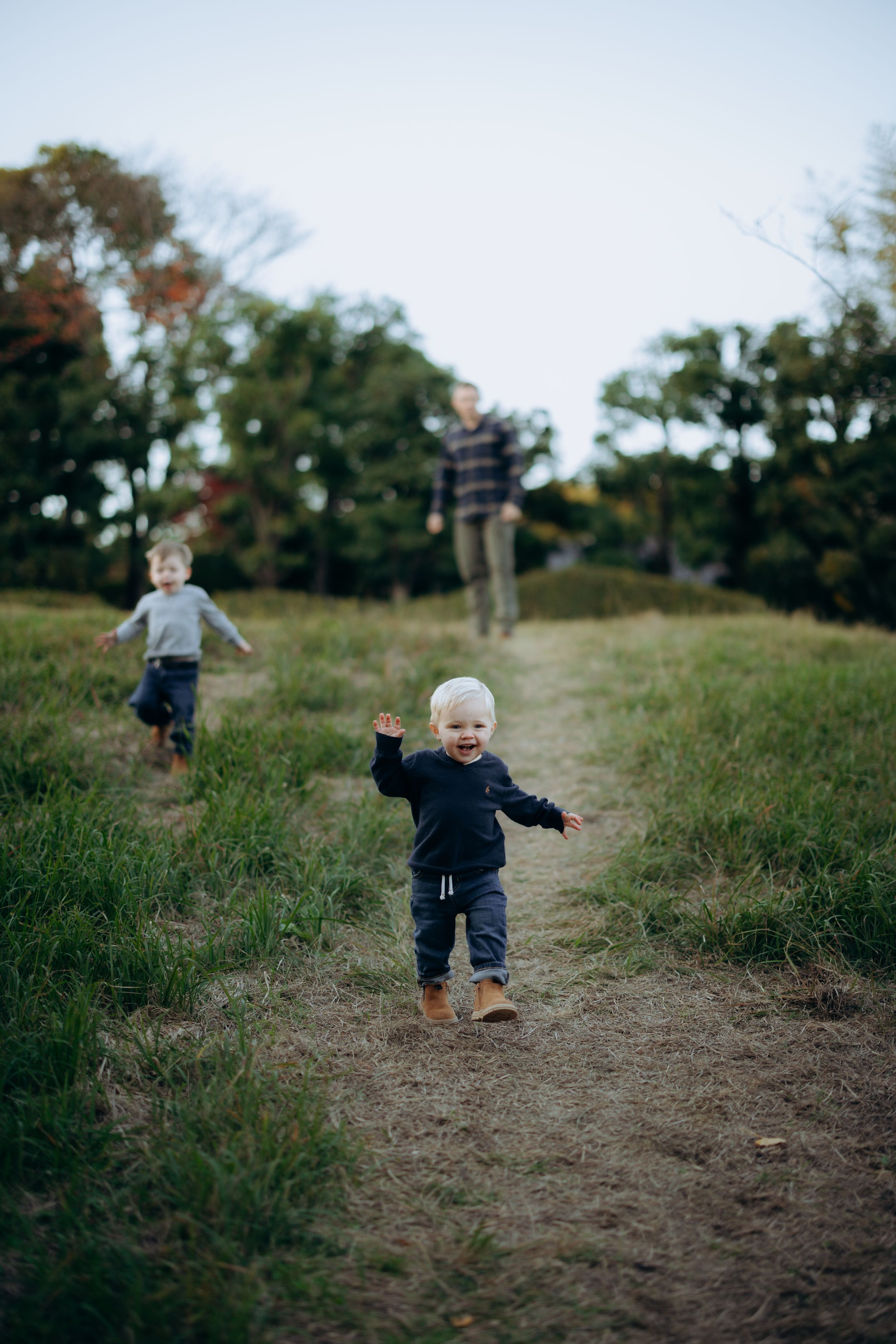 People walking along a dirt trail through grassy fields surrounded by trees on a cloudy day.