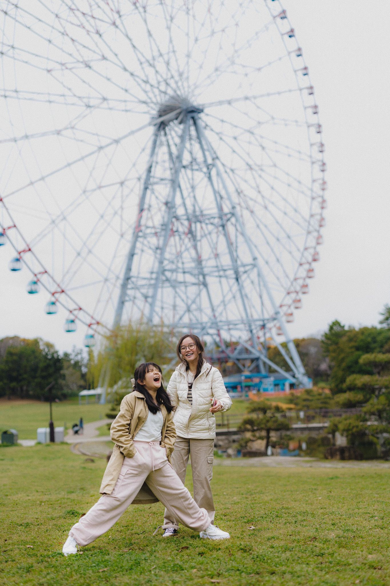 Two people strike fun poses on grass with a giant ferris wheel in the background.