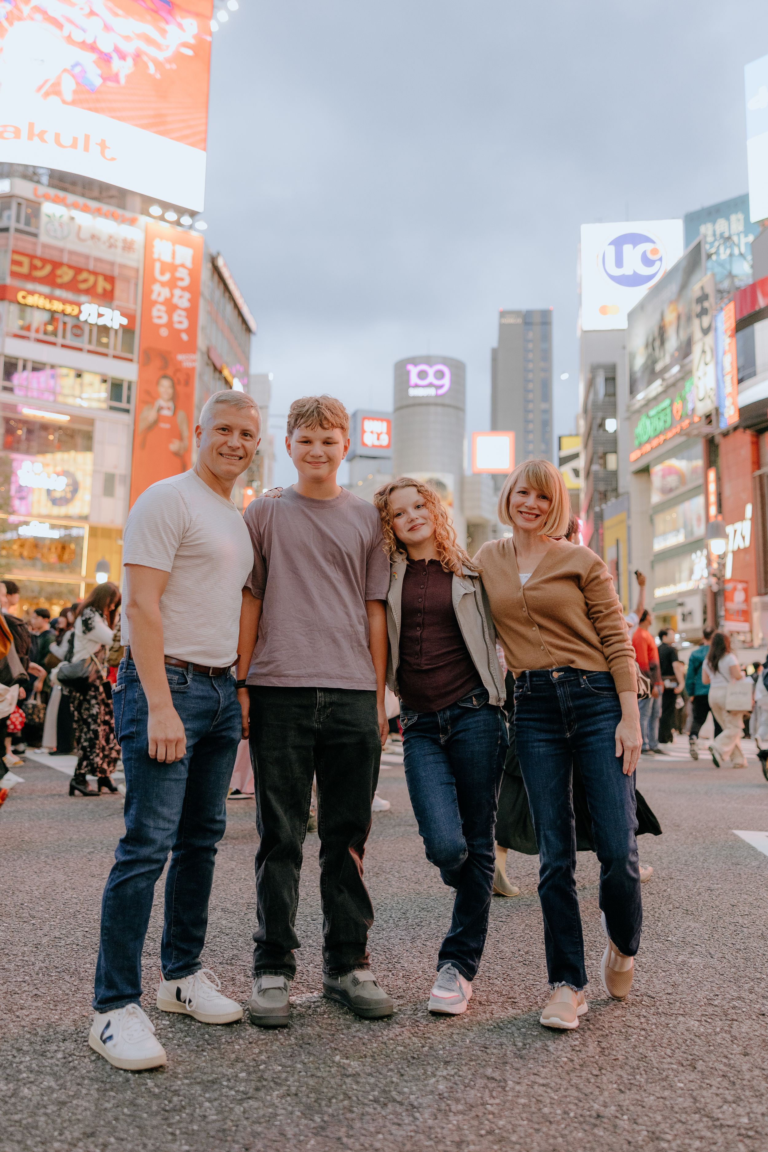Group poses together on busy city street with colorful neon signs and storefronts visible in Shibuya, Tokyo.