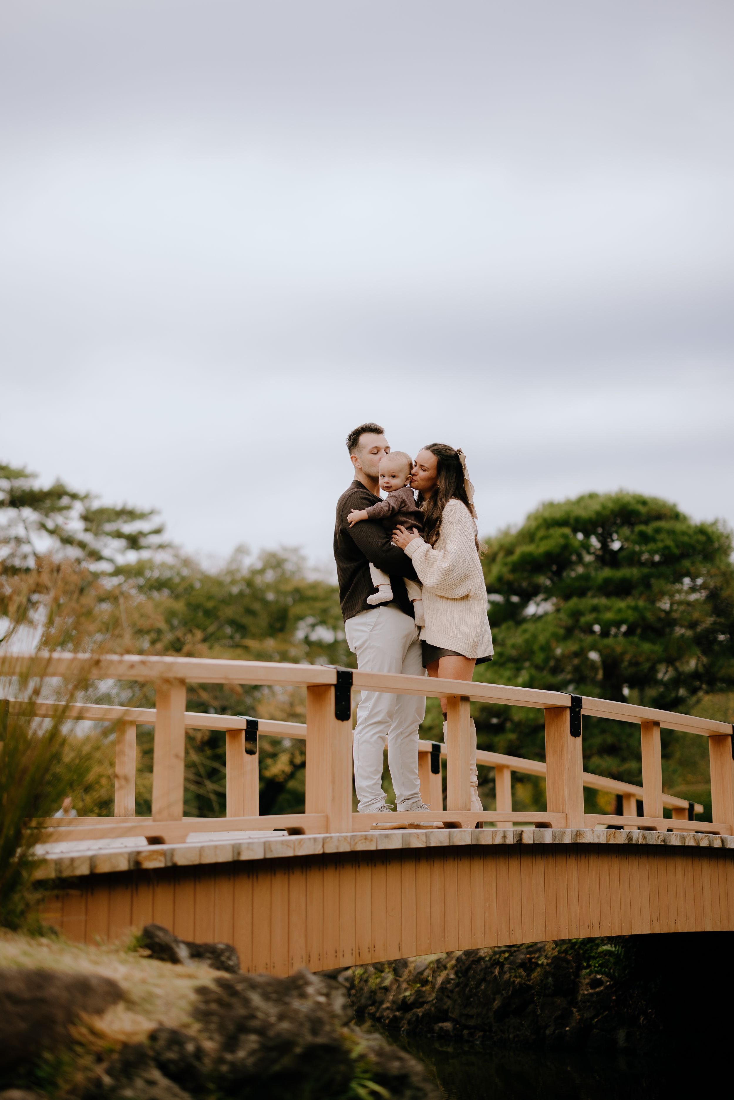 A couple shares a romantic moment on a wooden footbridge against a cloudy sky during a photoshoot.