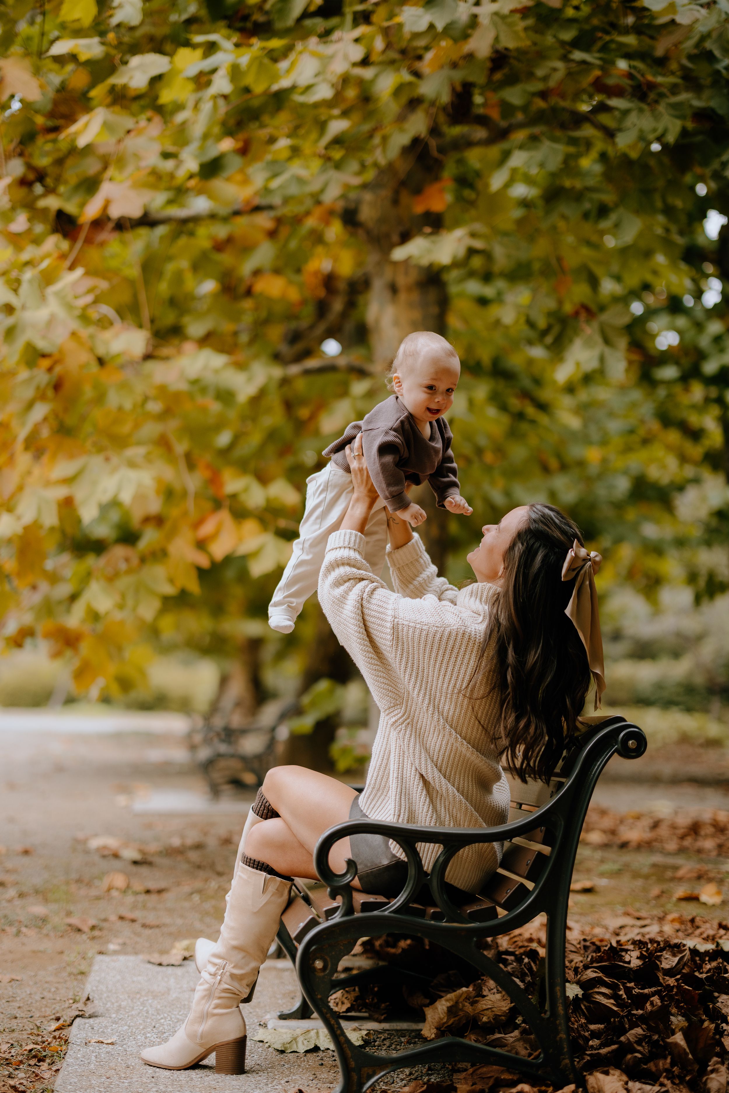 Person in cream sweater and boots sits on park bench surrounded by autumn foliage in scenic outdoor setting.