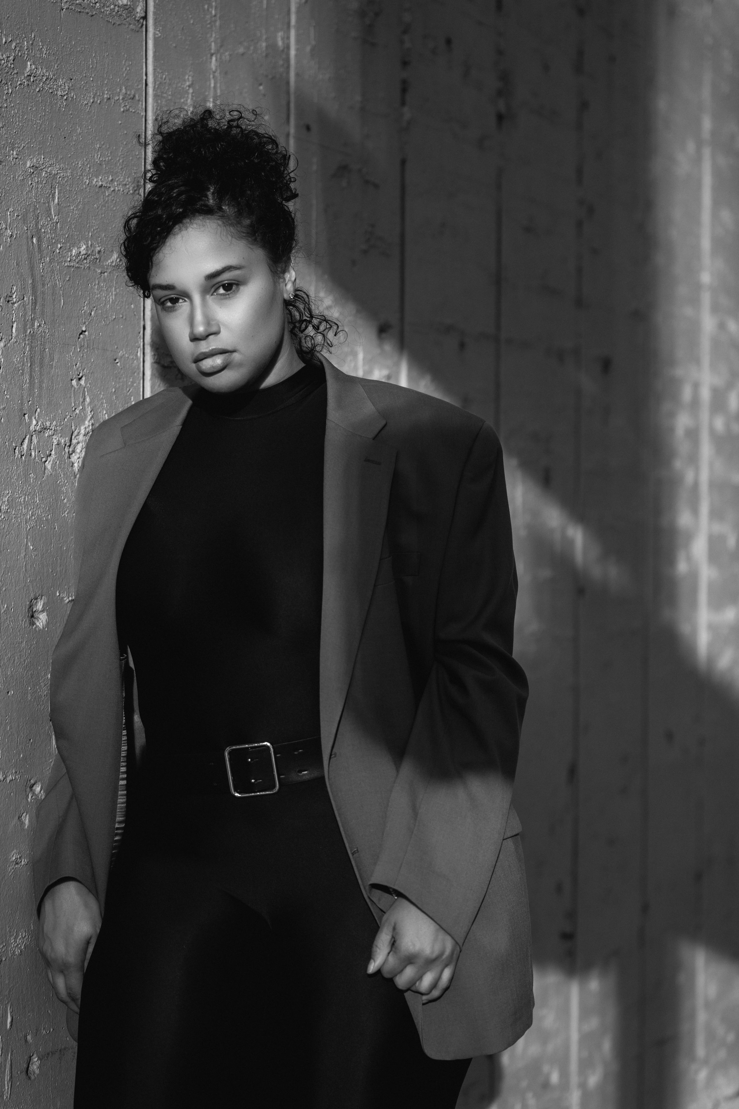 Black and white fashion portrait of a person in a blazer and turtleneck against a textured wall with dramatic lighting.