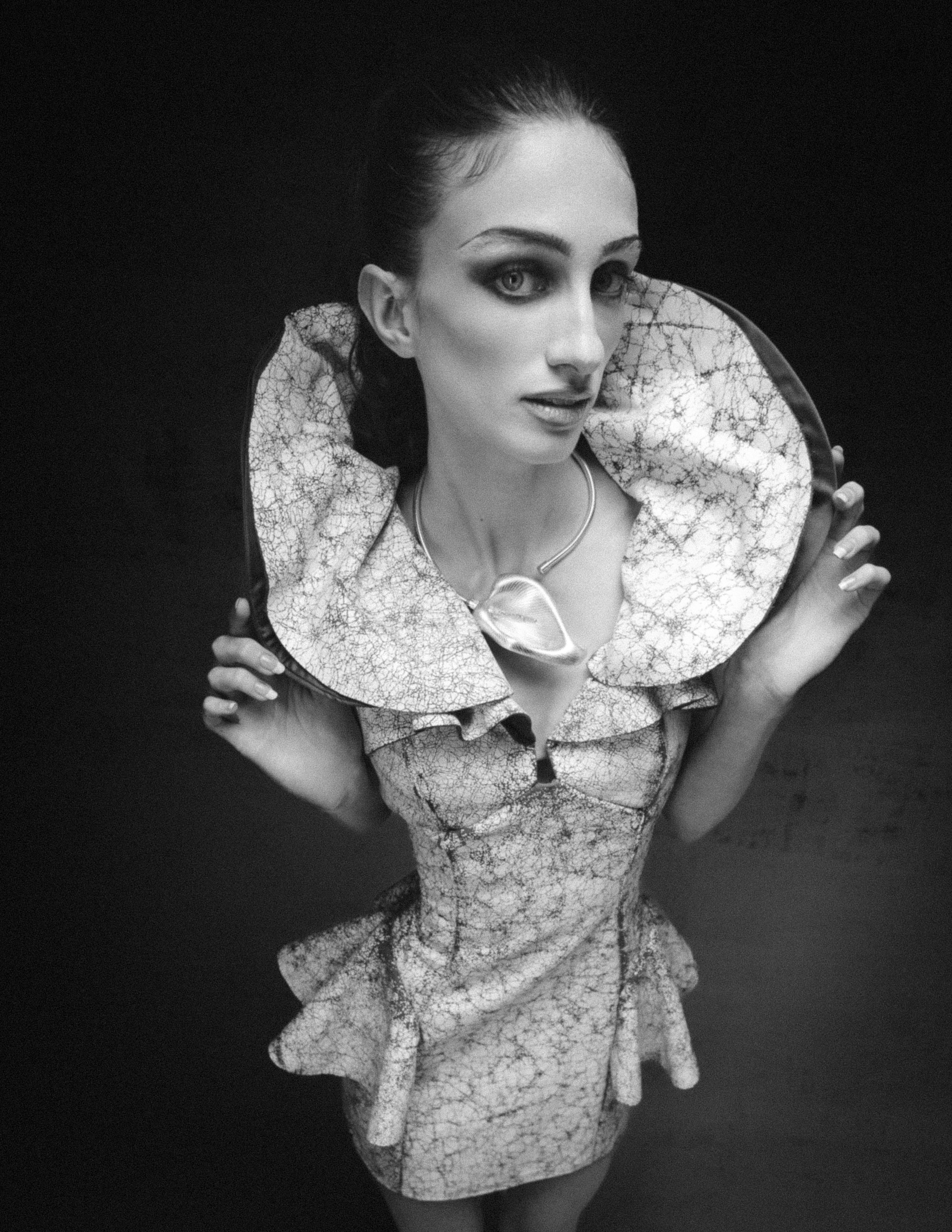 Black and white fashion photo of a model in an ornate ruffled collar dress, posing dramatically against a dark background.