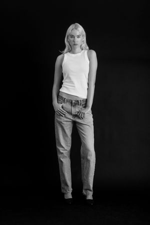 Black and white full-length portrait of a blonde woman in white tank top and jeans posing against dark background.