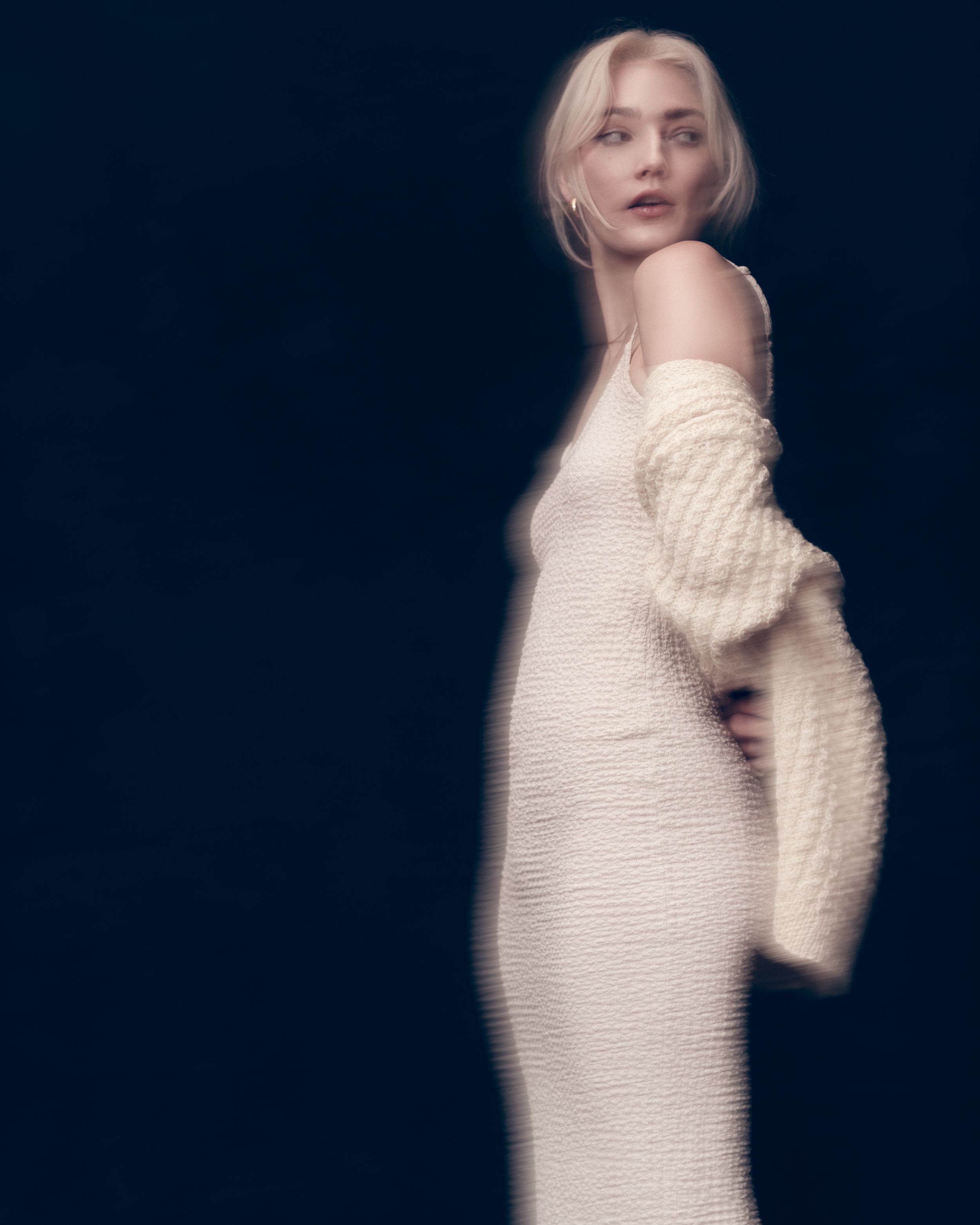 Fashion portrait on dark background shows figure in white knit dress with off-shoulder design in ethereal dreamy style.