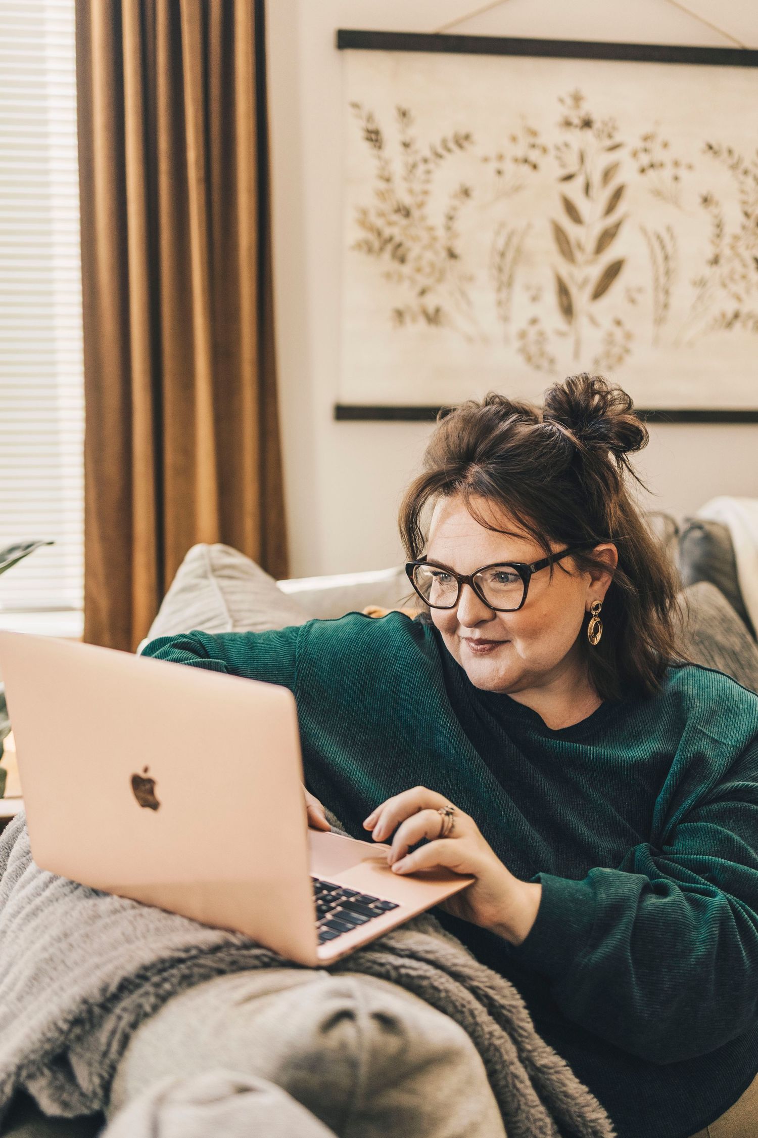 Woman with glasses in green sweater using Apple MacBook laptop on bed.