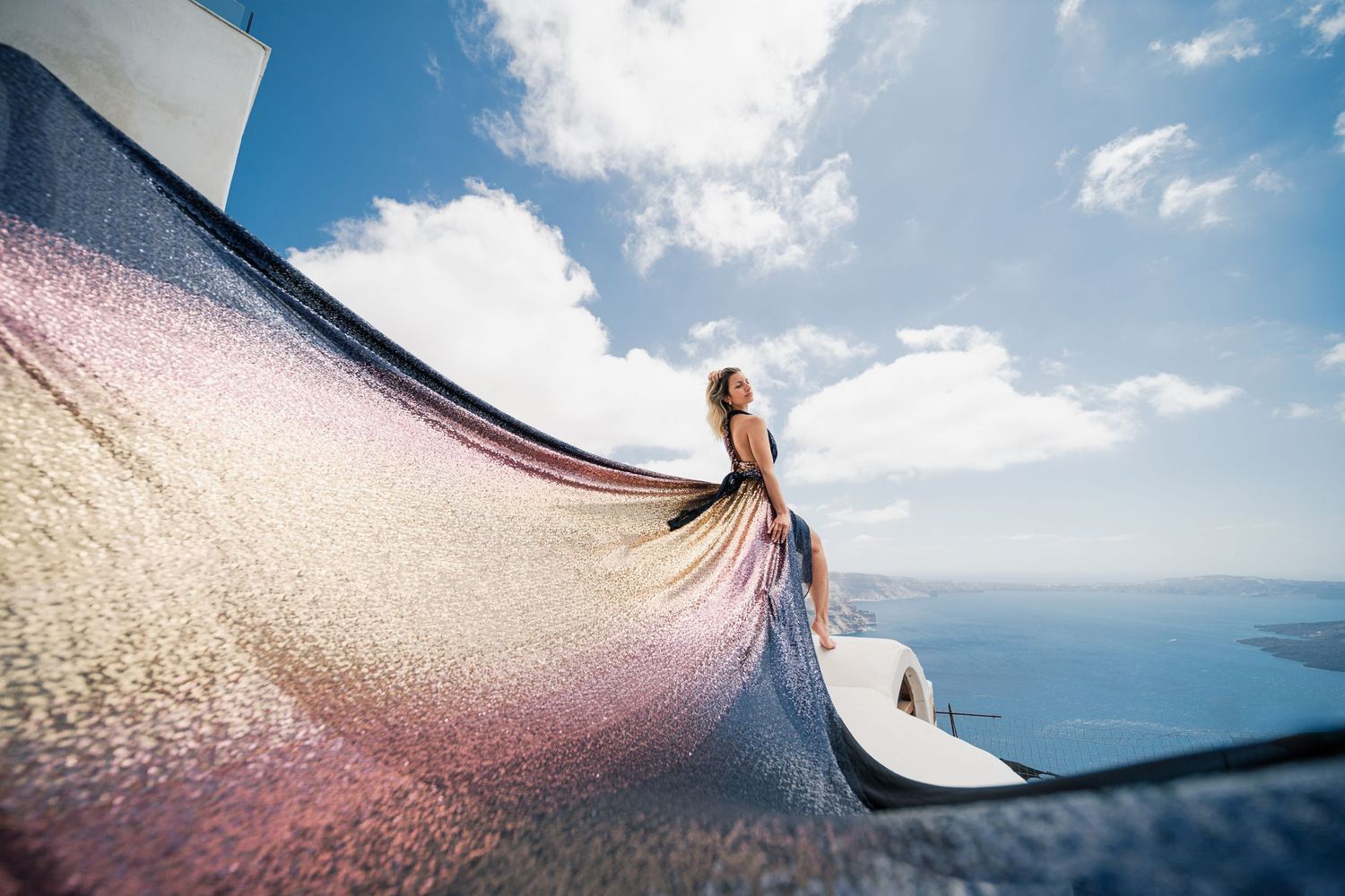 Shimmering ombre dress flows dramatically against classic Santorini white architecture and sea views.