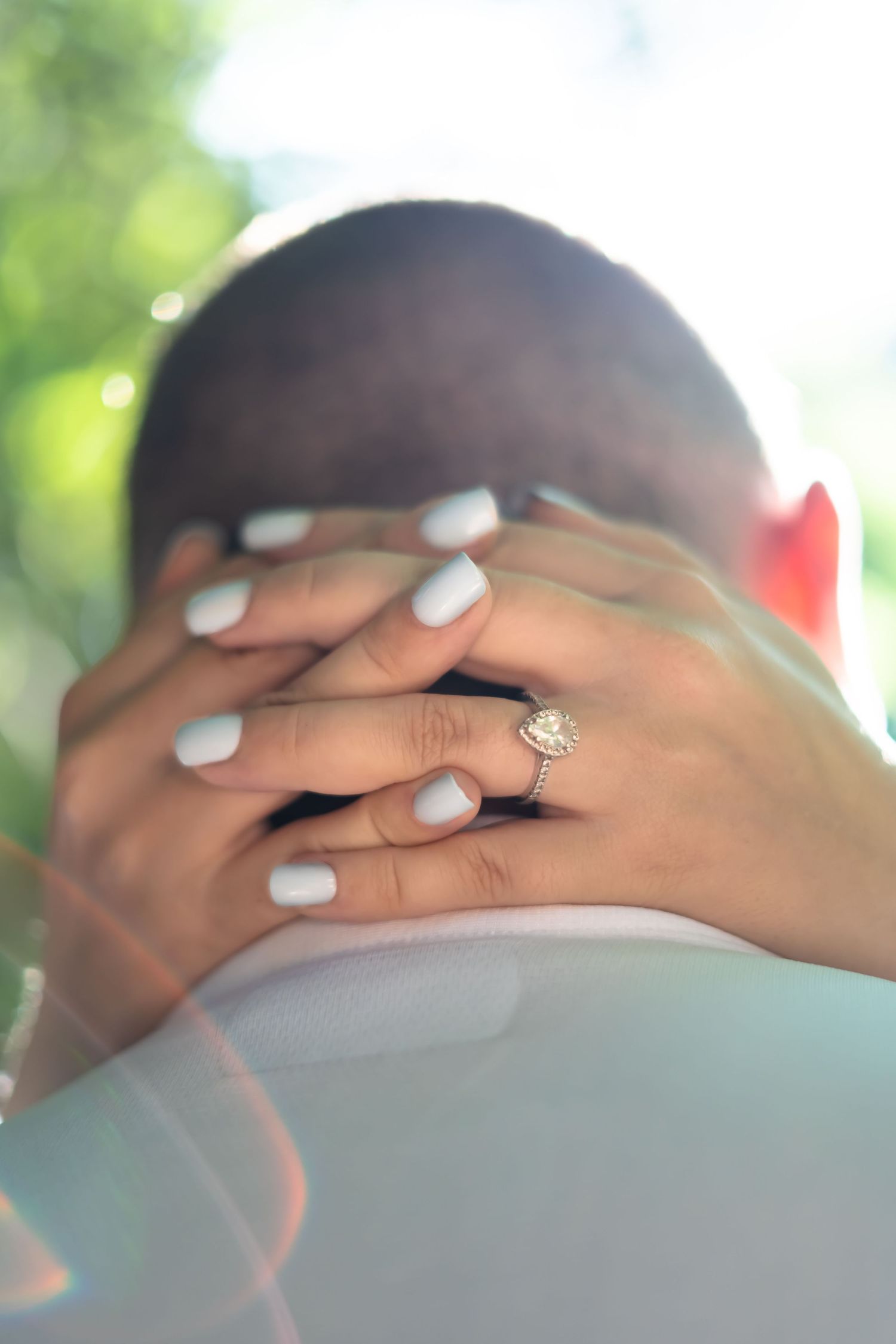Engagement ring detail shot with manicured hands against blurred background.