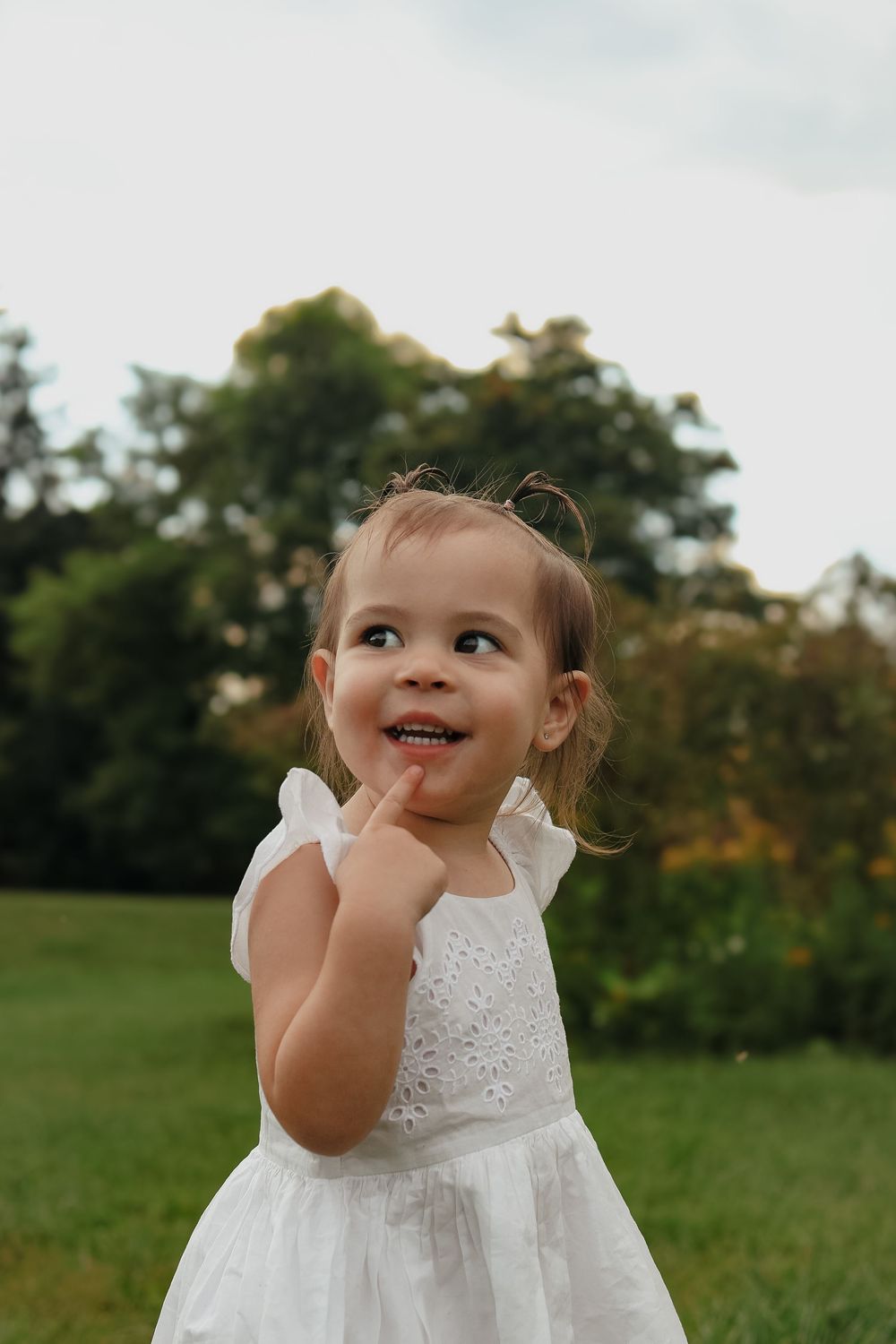 A toddler in a white lace dress plays joyfully on a grassy field during a summer day.