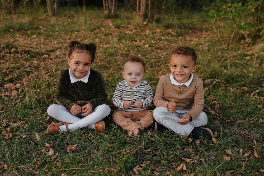 Three children sit together on fallen autumn leaves in a wooded outdoor setting.