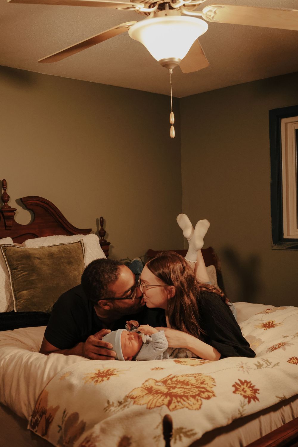 A couple lounging together on a floral bedspread beneath a ceiling fan in a cozy bedroom.