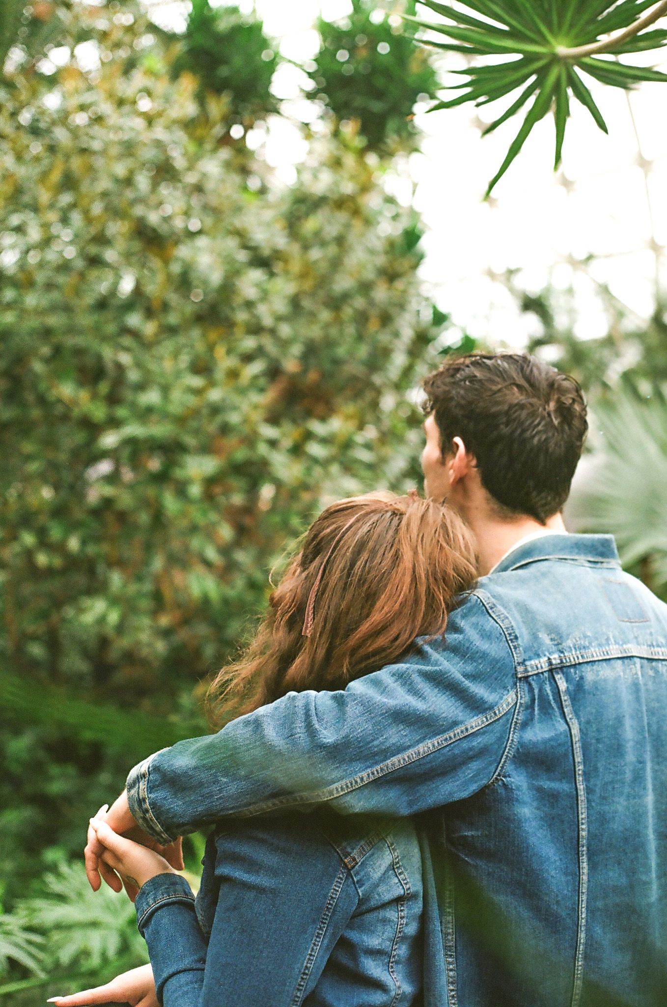 Couple in denim jackets embrace while viewing lush garden scenery.