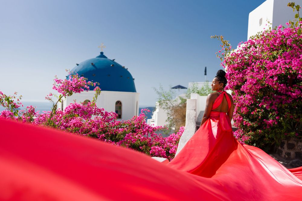 Flowing red fabric creates dramatic movement against classic Santorini architecture and pink flowers.