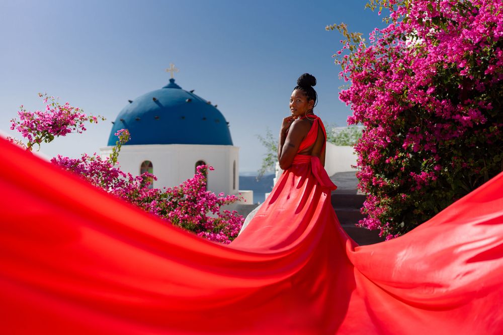 Red dress fabric swirls dramatically against blue-domed church and pink bougainvillea in Santorini.