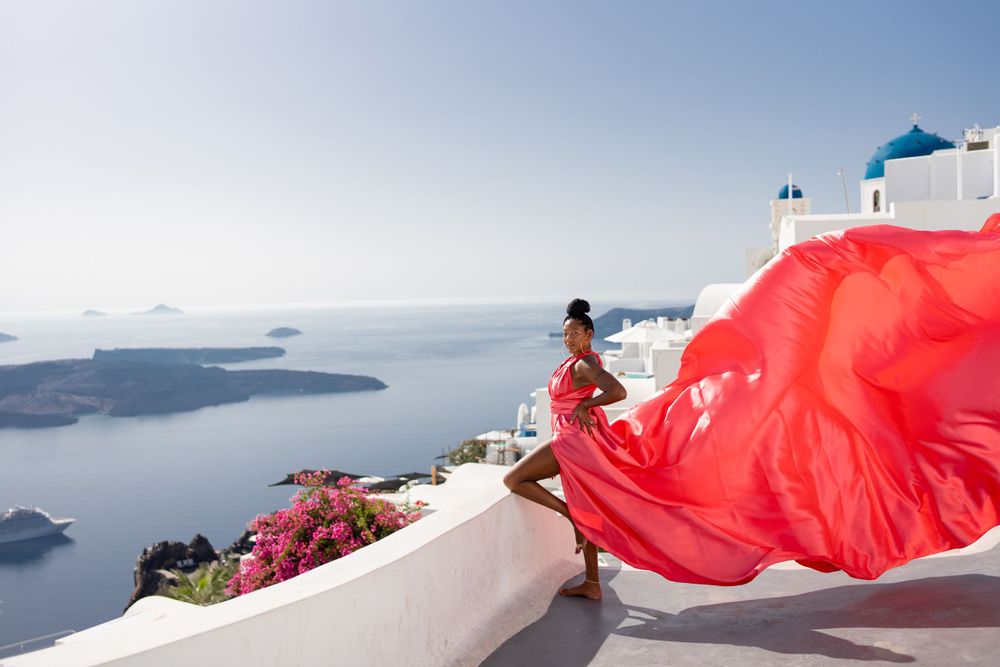 Red fabric billows dramatically on a white terrace overlooking Santorini's caldera and blue sea.