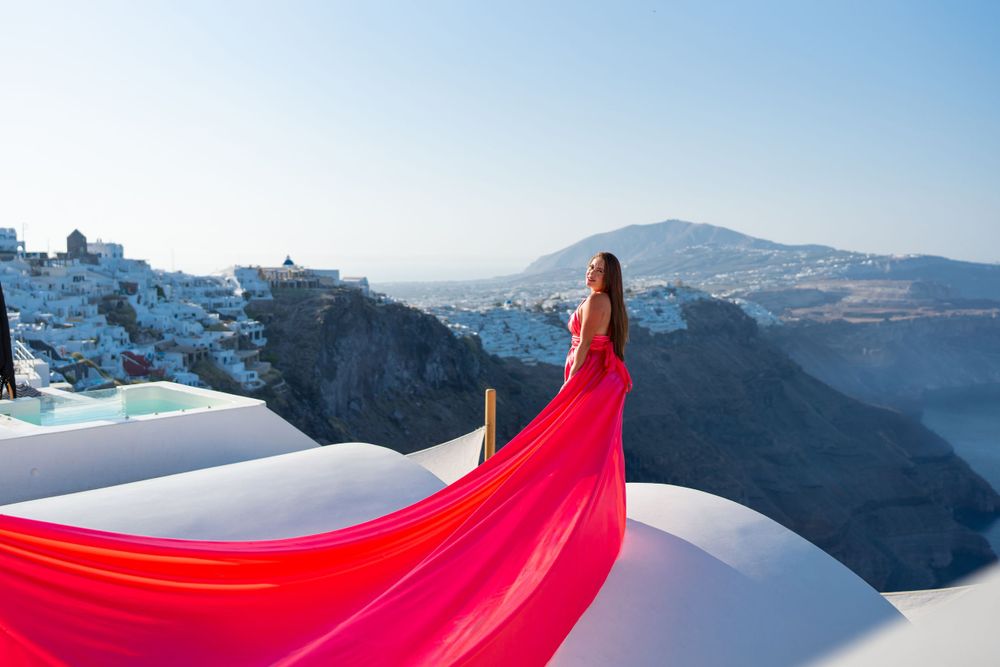 Dramatic red dress flows in the wind on white terrace overlooking Santorini's volcanic caldera.