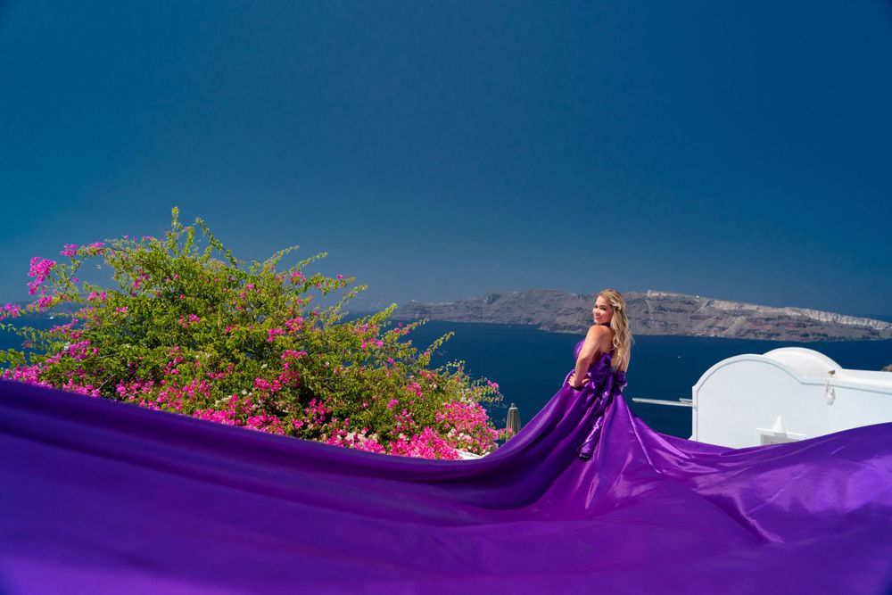 A flowing purple gown contrasts with pink flowers and white architecture in Santorini.