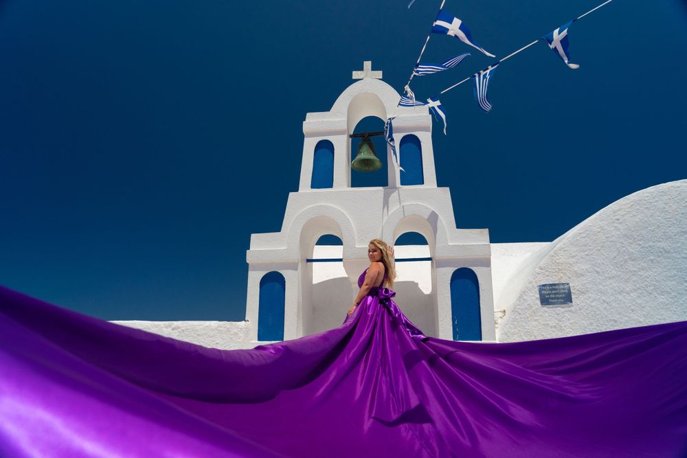 Flowing purple gown against white bell tower and blue sky in Santorini.