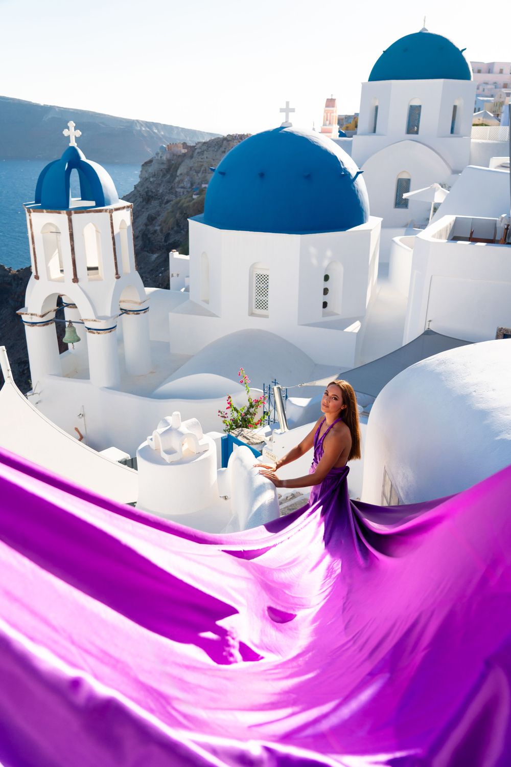 Purple fabric swirls dramatically against white architecture and blue domes in Santorini.
