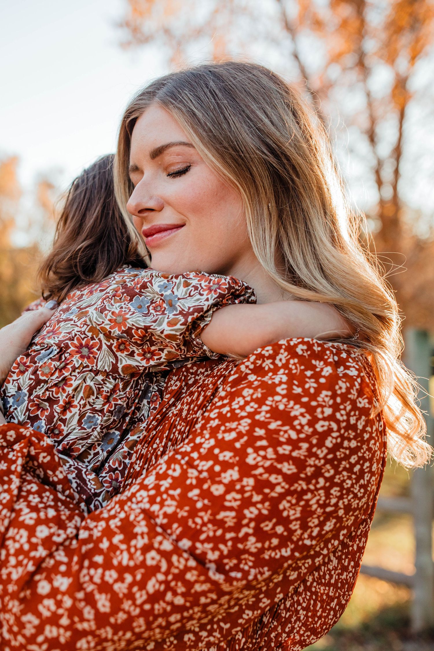 Woman wearing orange floral dress poses in autumn setting with golden sunlight.