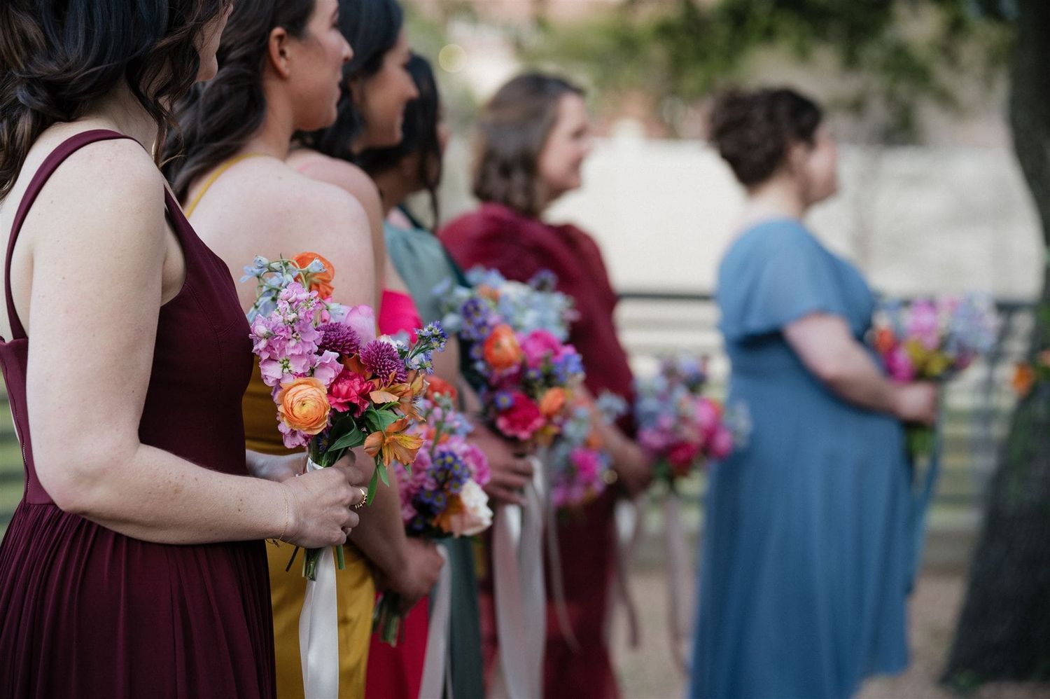 A line of bridesmaids in burgundy and blue dresses holding vibrant multicolored floral bouquets at an outdoor wedding.