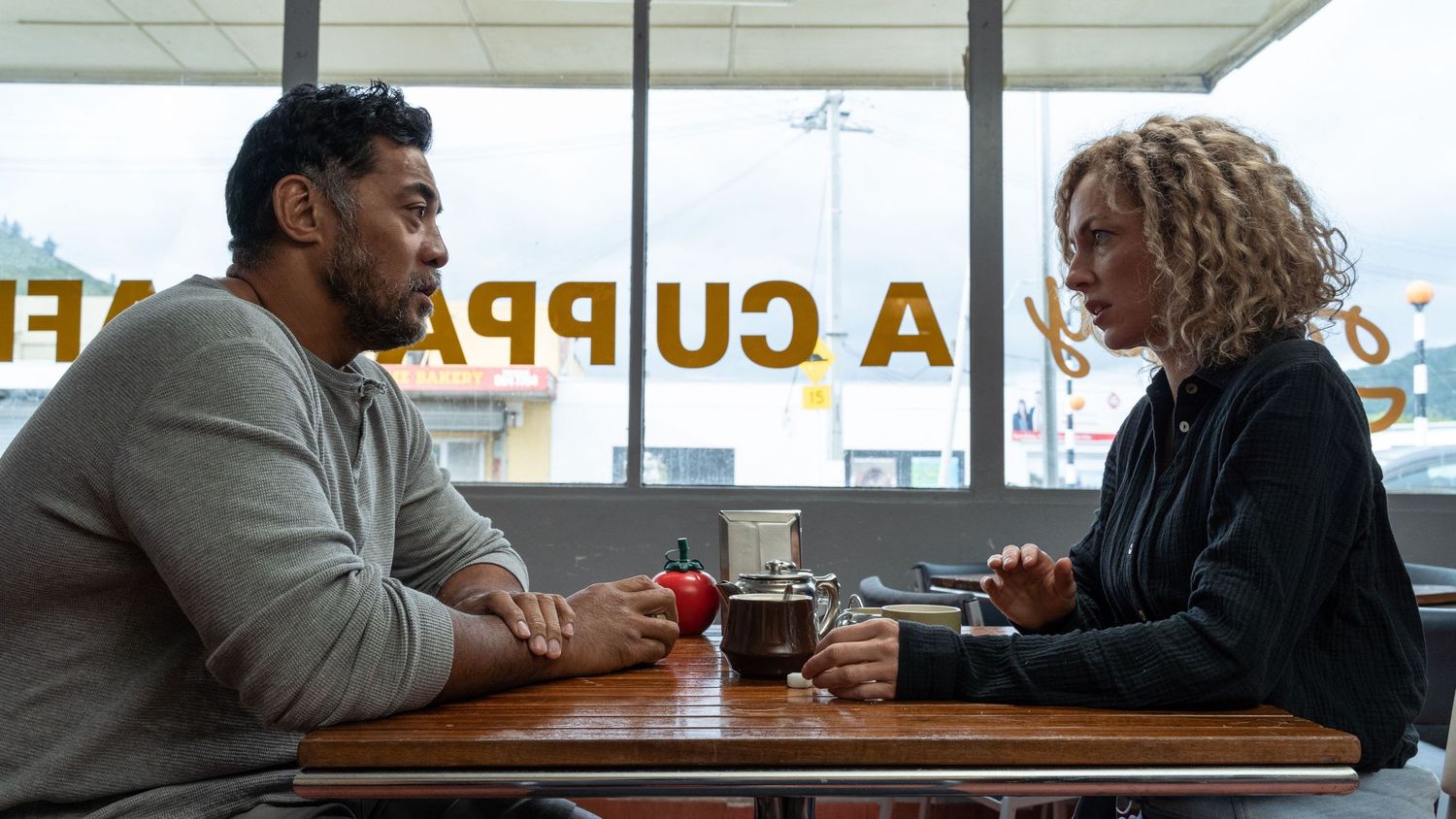 Robbie Magasiva and Loren Taylor in character sitting at a cafe table across from each other. A still from the movie