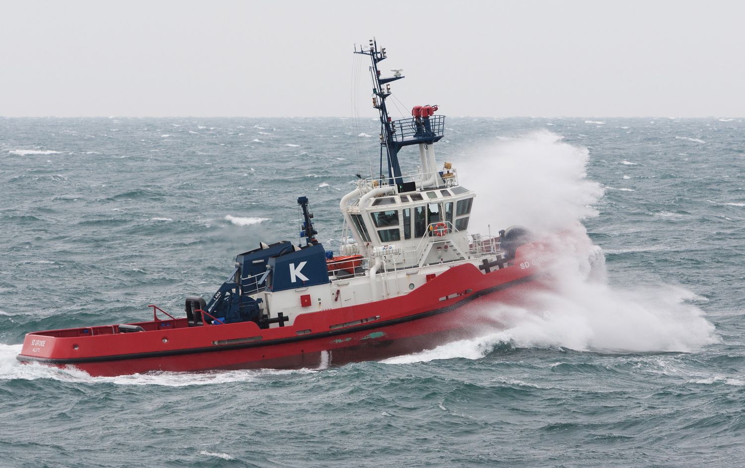 Red and white tugboat powering through rough seas with spray flying in strong winds.