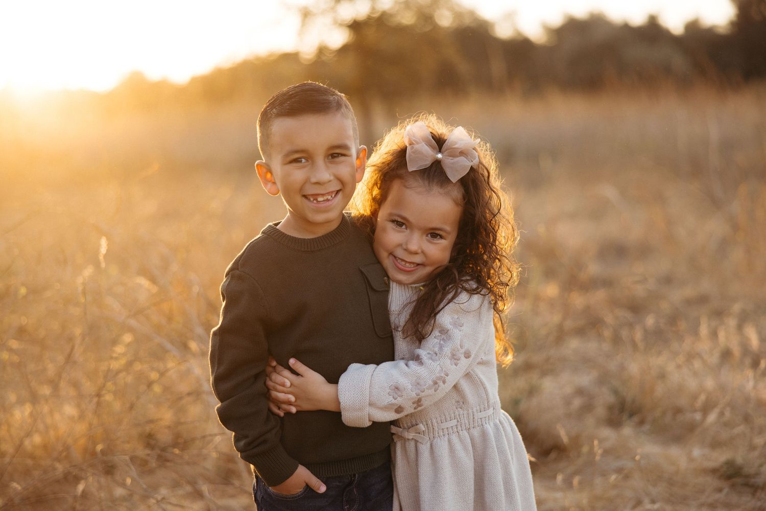 Two young children embrace while standing in a sunlit field at golden hour.