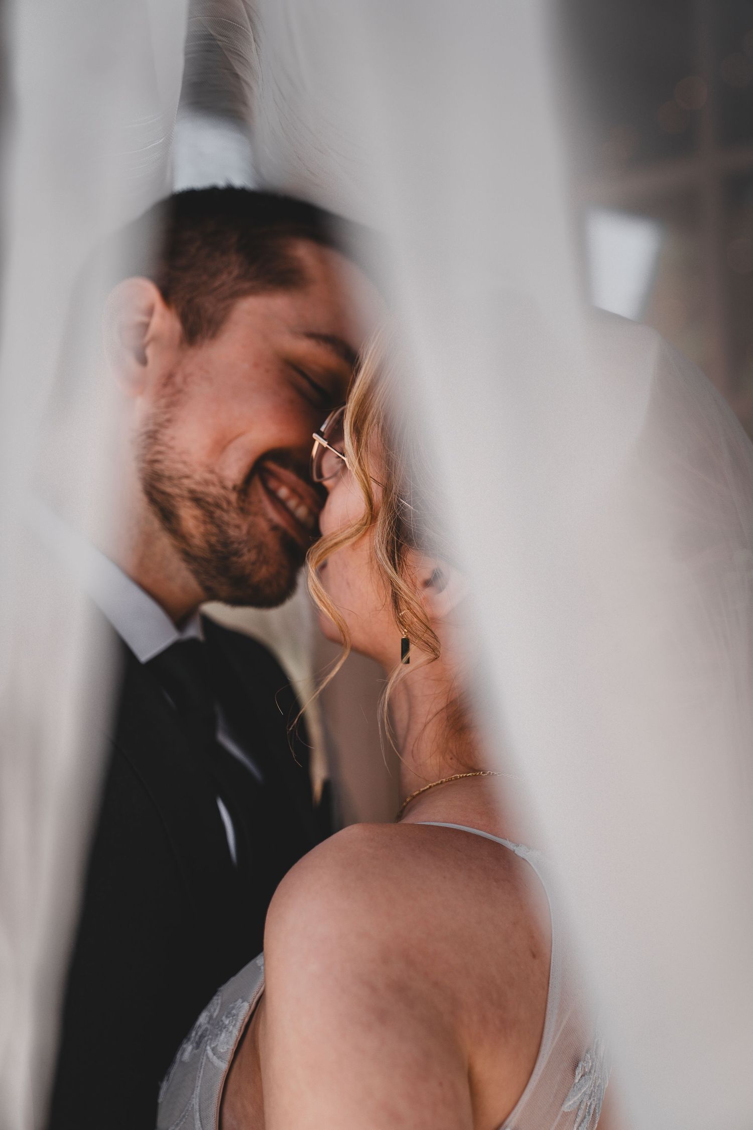 Intimate romantic wedding moment captured through sheer veil as couple shares tender moment in soft lighting.