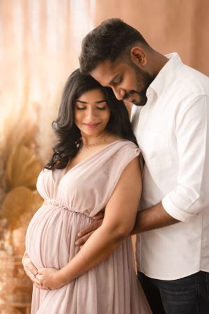 Intimate maternity portrait of couple in white and pink clothing against warm backdrop.